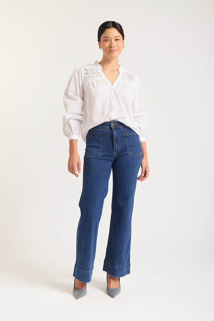 Woman against white background in white shirt with lace details at the shoulder.