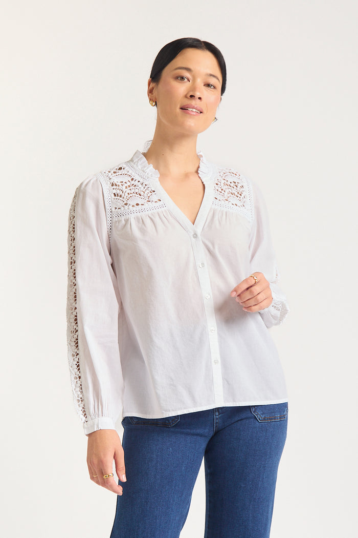 Woman against white background in white shirt with lace details at the shoulder.