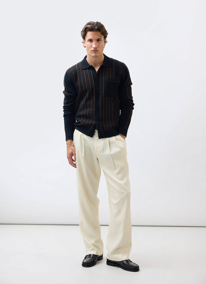 A model wears the Percival Appendix Long Sleeve Knitted Shirt in black and brown with cream trousers and black loafers.