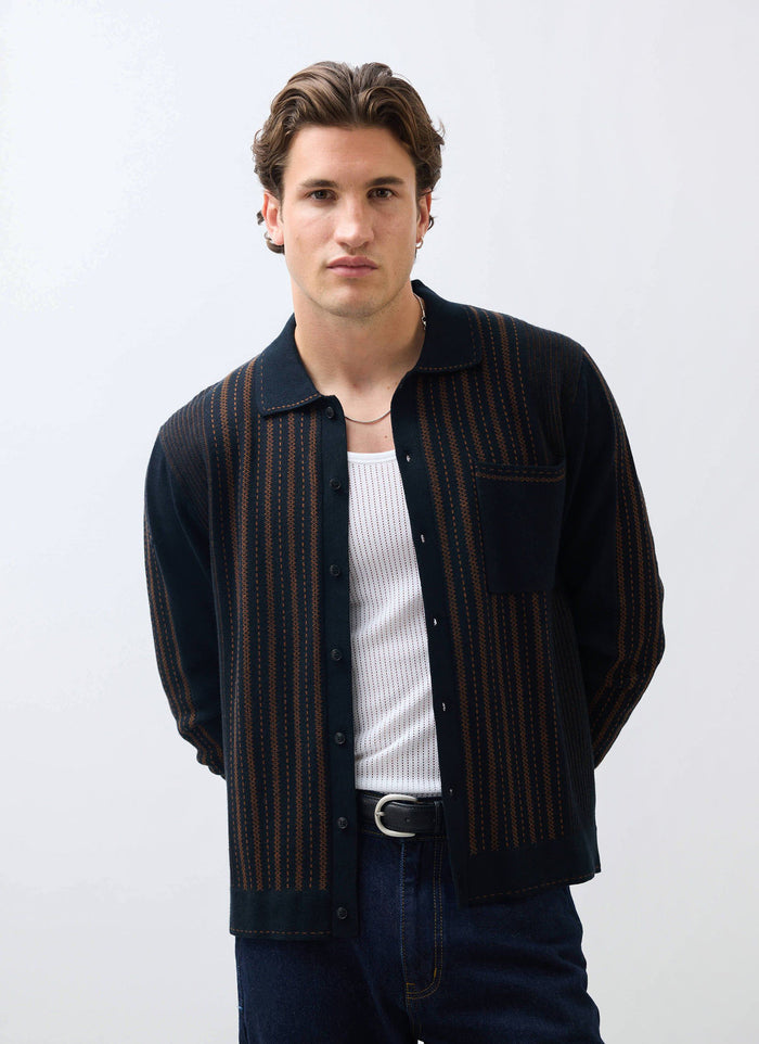 A model wears the Percival Appendix Knitted Shirt in black and brown, styled open over a white vest and jeans.