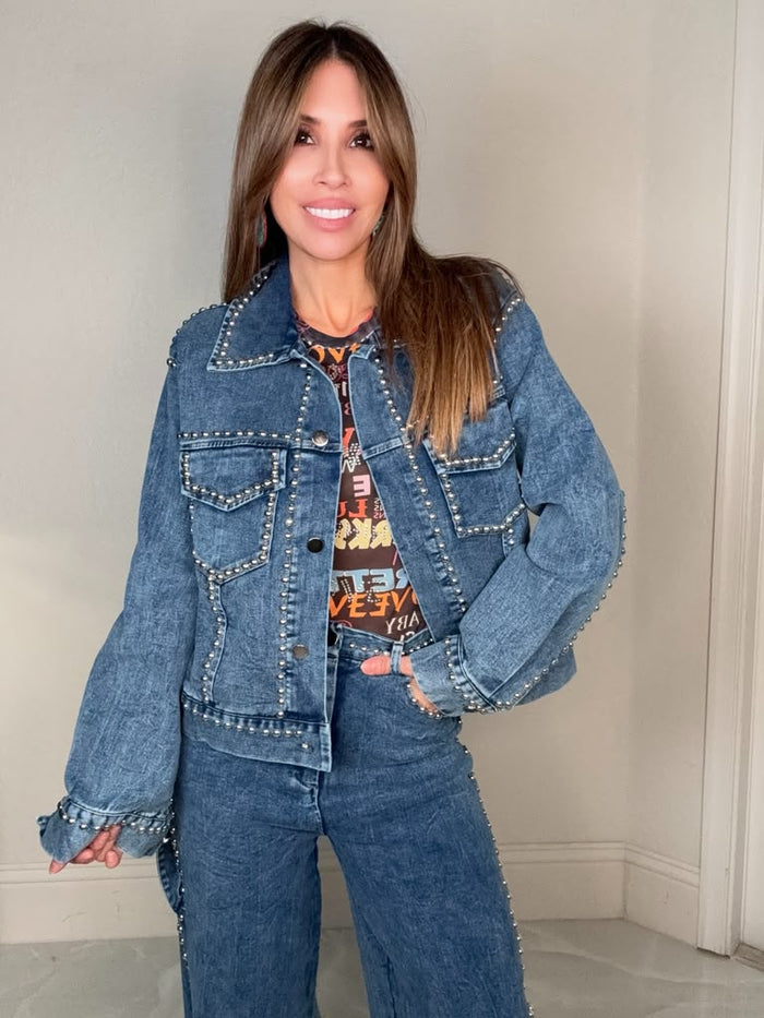Blue denim jacket with metal stud embellishments along the seams and front pockets in a structured button-front silhouette.