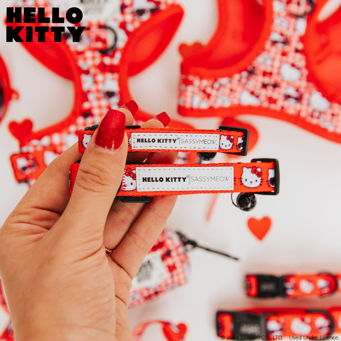 A hand with red-painted nails holds two Sassy Woof Cat Collar - Hello Kitty® Red Classic tags next to matching red and white Sanrio accessories and small red hearts. The Hello Kitty logo is in the top left corner. Ideal for any adjustable collar.