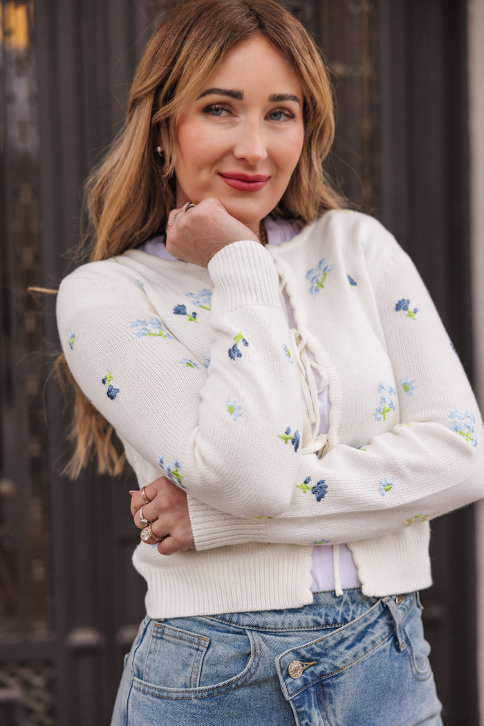 A woman with long, light brown hair wears the Ivy City Co Della Sweater in Cream and blue jeans, standing outdoors with her hand on her chin and smiling softly at the camera.
