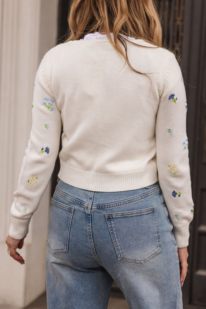A woman with long, wavy hair stands with her back to the camera, wearing Ivy City Co’s Della Sweater in Cream—adorned with colorful embroidered flowers on the sleeves and back—and light blue high-waisted jeans.