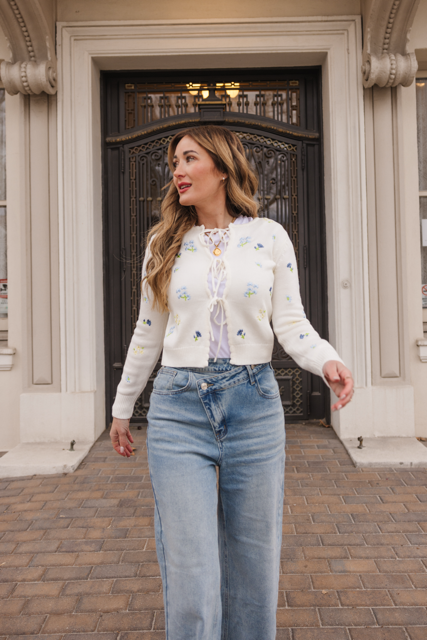 A woman with long, wavy hair wearing the Della Sweater in Cream by Ivy City Co and blue jeans walks confidently on a brick path in front of an ornate black door and white building facade.