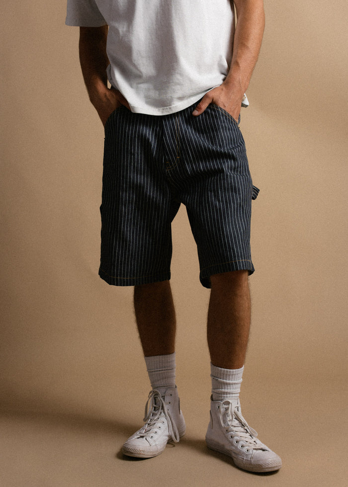 Man in white shirt and striped shorts stands with hands in pockets, wearing white sneakers and socks against a beige background.