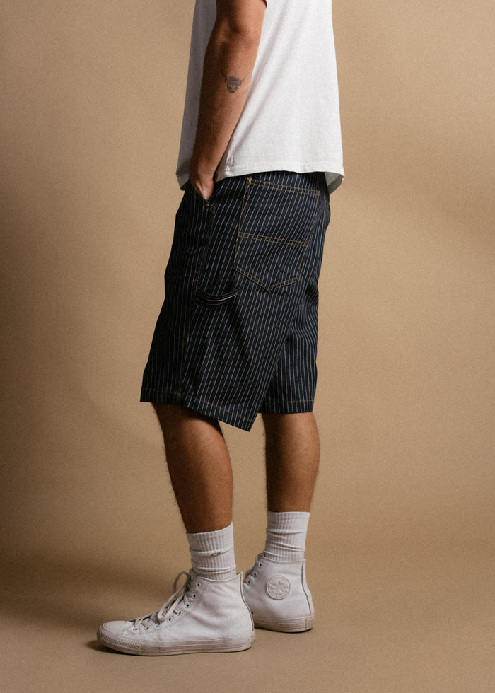Man in white sneakers and striped shorts stands against a beige background, showcasing casual style with a tattoo on his arm.