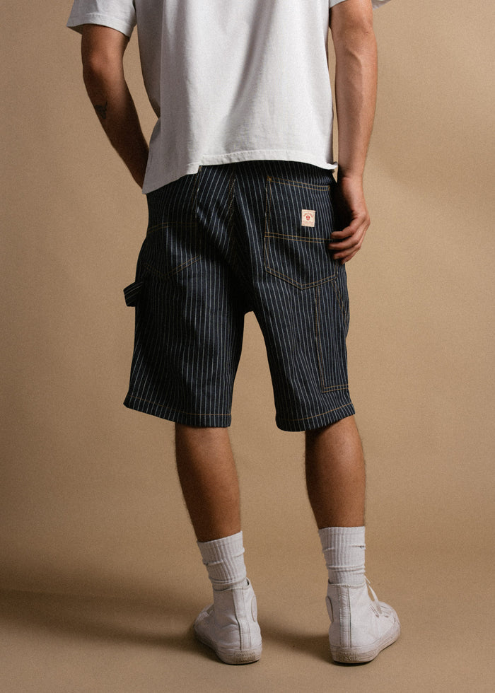 Person in a white shirt and striped shorts stands against a beige background, wearing white socks and sneakers.