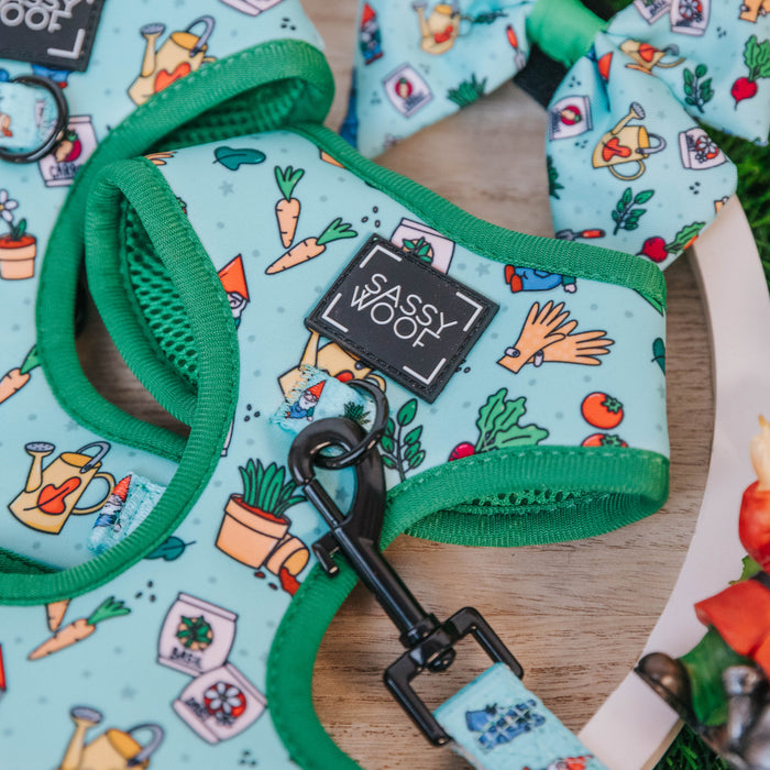 A close-up of the Sassy Woof Dog Adjustable Harness - Chillin with my Gnomies in light blue with green trim, featuring colorful gardening-themed prints. The matching no-pull leash and bowtie are shown alongside the harness.