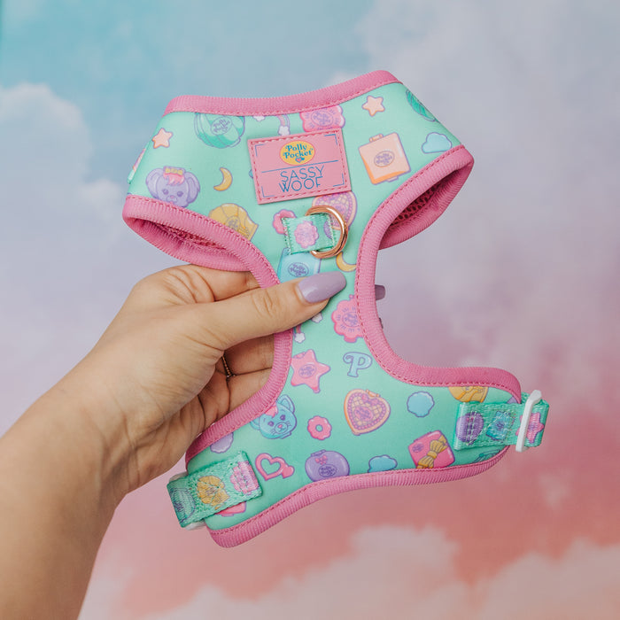 A hand holds the Sassy Woof Dog Adjustable Harness - Polly Pocket™ in pastel blue and pink, featuring colorful cartoon icons like hearts, stars, and paw prints, set against a soft, cloudy gradient background.