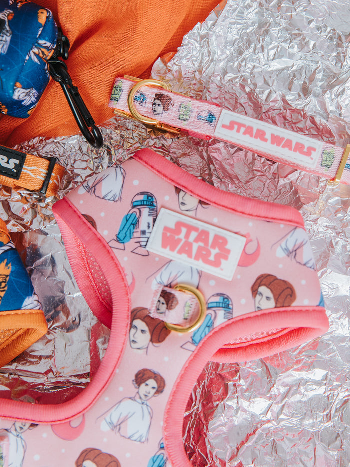 The Sassy Woof Dog Adjustable Harness - STAR WARS™ Princess Leia, featuring a pink Rebel Alliance design, is paired with a matching leash and displayed on crinkled silver foil and orange fabric.