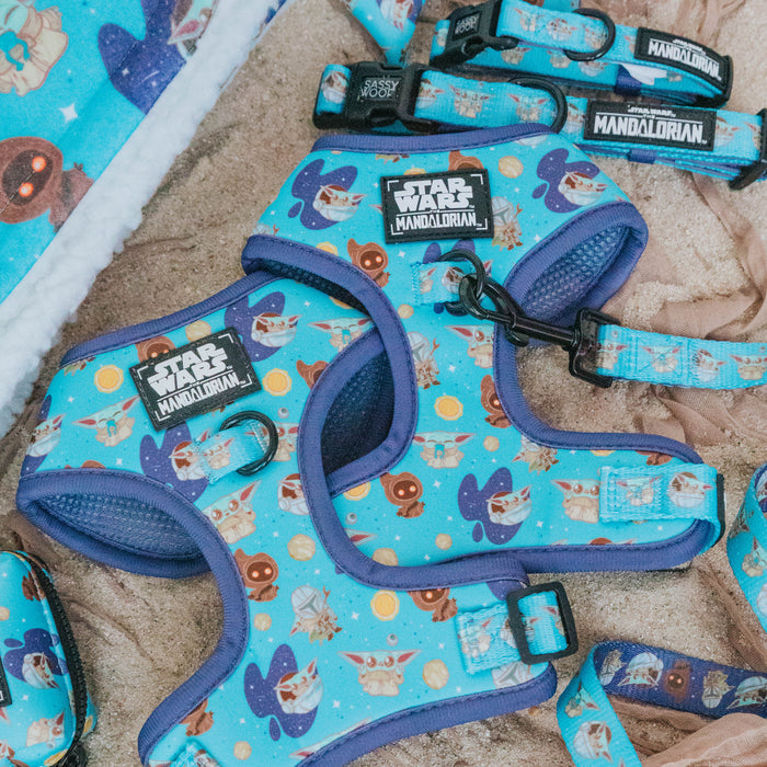 Two Sassy Woof STAR WARS™ The Mandalorian™ Dog Adjustable Harnesses with cartoon Baby Yoda and space designs lie on sand beside a soft blanket.