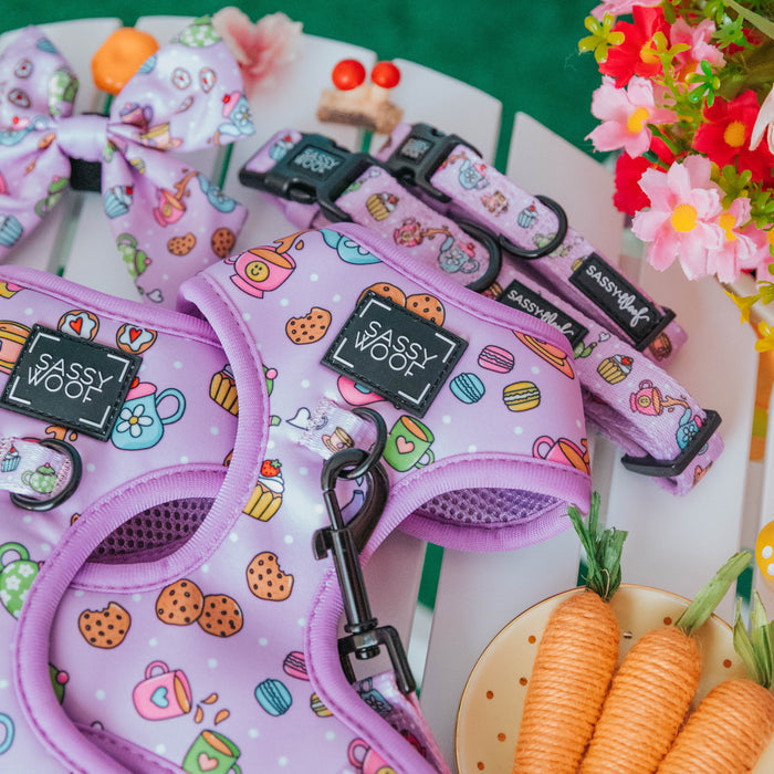 The Sassy Woof Dog Adjustable Harness – Tea Pawty is a pastel purple set with cute snack designs, shown with a collar and leash on a white table near toy carrots, a floral bow, and artificial flowers for stylish puppy protection.
