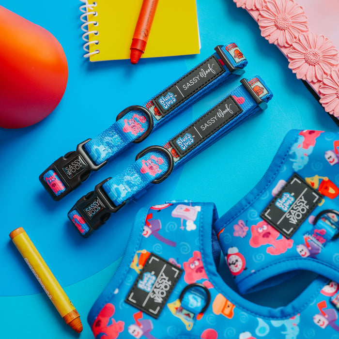 Two Sassy Woof Dog Collar - Blue's Clues™ accessories, featuring colorful cartoon sea creatures, are displayed on a blue surface near crayons, a yellow notebook, and a pink floral item.