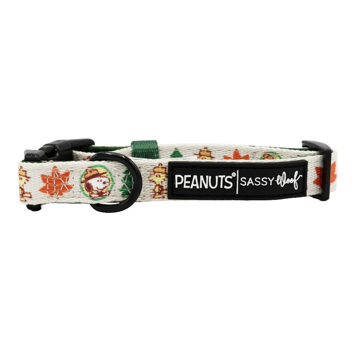 The Sassy Woof Dog Collar - Peanuts™ Beagle Scouts is a beige adjustable collar with autumnal Snoopy and leaf designs, a black buckle and D-ring, plus a black patch reading "PEANUTS | SASSY WOOF" in white text.