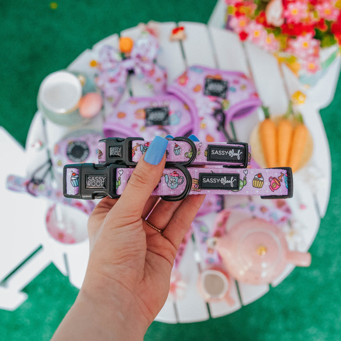 A hand with blue-painted nails holds two Sassy Woof Dog Collar - Tea Pawty collars above a white table set with tea items, flowers, and pet accessories; the purple collars feature cupcake and ice cream designs plus a sturdy D-ring.