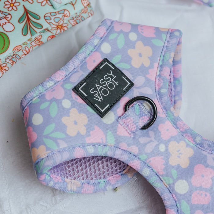 A close-up of the pastel purple Dog Four Piece Bundle - Fleur de Sass by Sassy Woof, featuring a floral pattern and black Sassy Woof label. The harness is on a white surface beside colorful fabric.