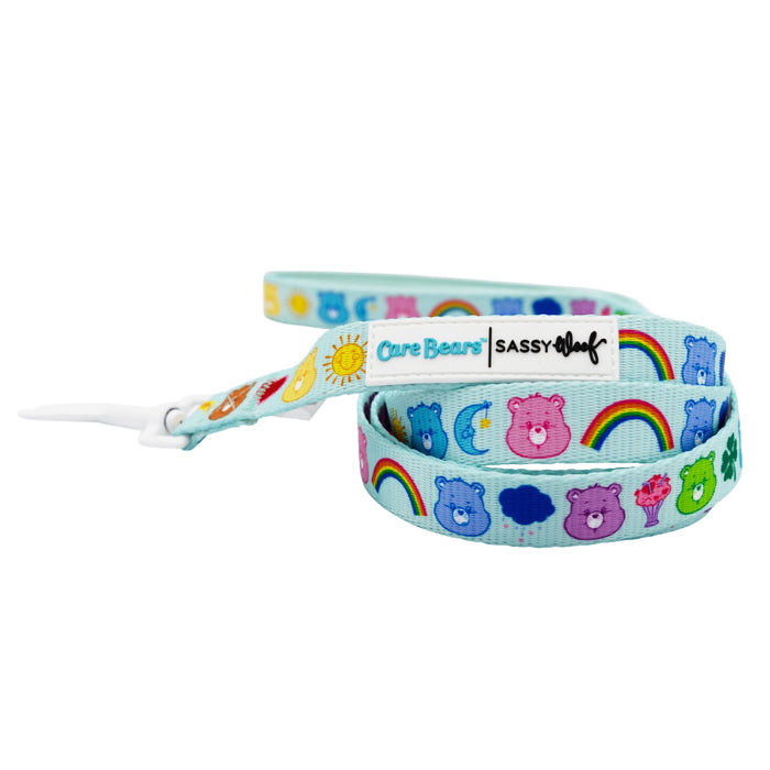 The Dog Leash - Care Bears™ Care-a-Lot by Sassy Woof features blue fabric with colorful Care Bears faces, rainbows, suns, moons, stars, and Care Bears and Sassy Woof logos on a white tag.