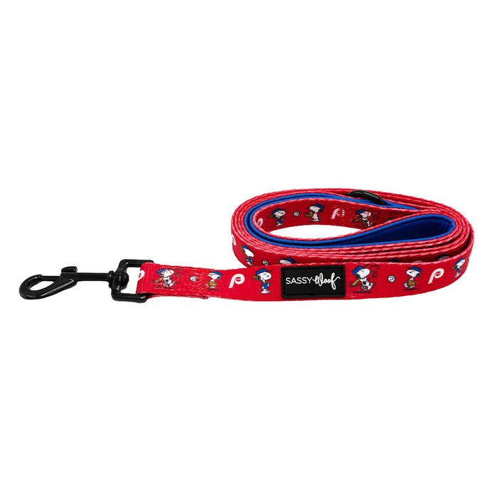 The Sassy Woof Dog Leash - MLB™ x Peanuts™ Philadelphia Phillies™ features red webbing with Snoopy and baseball graphics, a blue inner lining, a sturdy black metal clip, and an official Sassy Woof label—ideal for Phillies fans and their pets.
