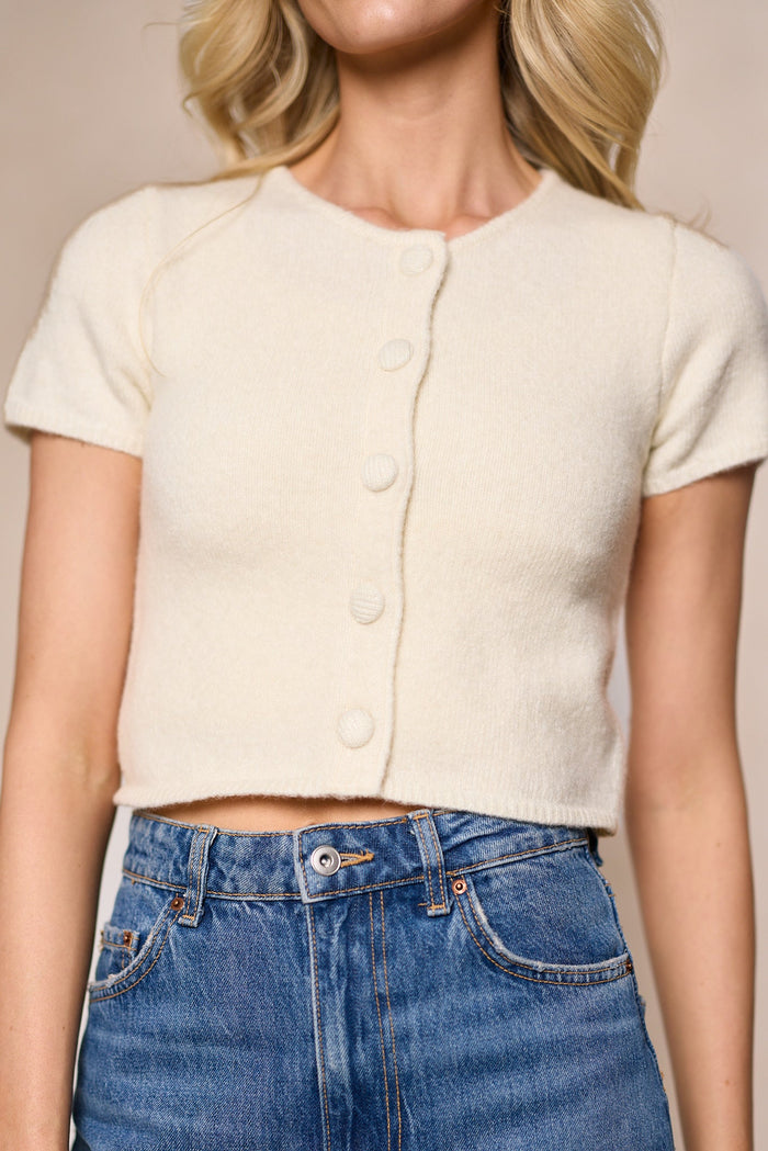A woman models the Elowen Cardigan in Ivory by Ivy City Co, styled as a cropped short-sleeve button-front top with high-waisted blue jeans. Only her torso and part of her wavy blonde hair are visible; her face is not shown.