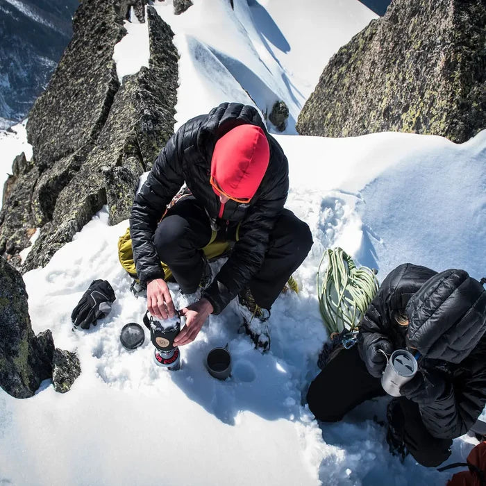 Person in a snowy mountain setting preparing a camping stove whilst wearing a Sub Zero down jacket and red balaclava.