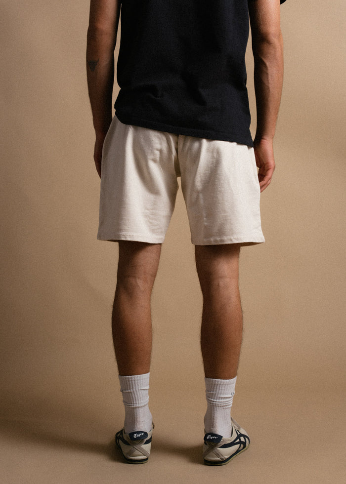 Man in black shirt and beige shorts stands facing away, wearing white socks and sneakers, against a plain beige background.