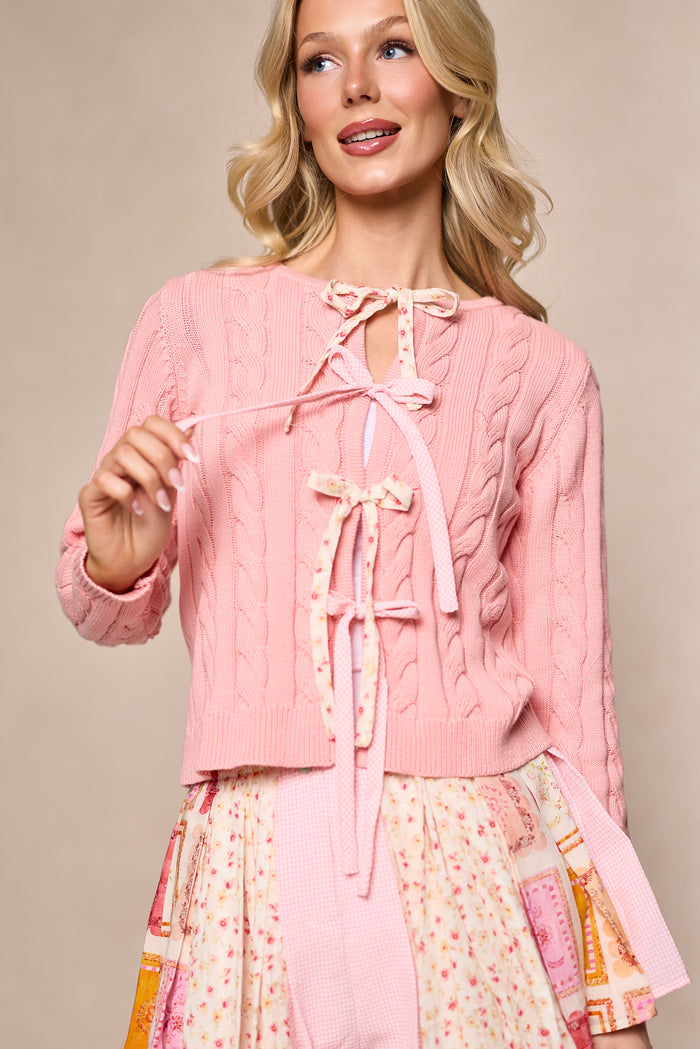 A smiling woman models the Ivy City Co Imani Sweater in Blush, a light pink 100% cotton cable-knit cardigan with three front ribbon bows, paired with a flowy floral skirt. She holds one ribbon tie against a beige background.