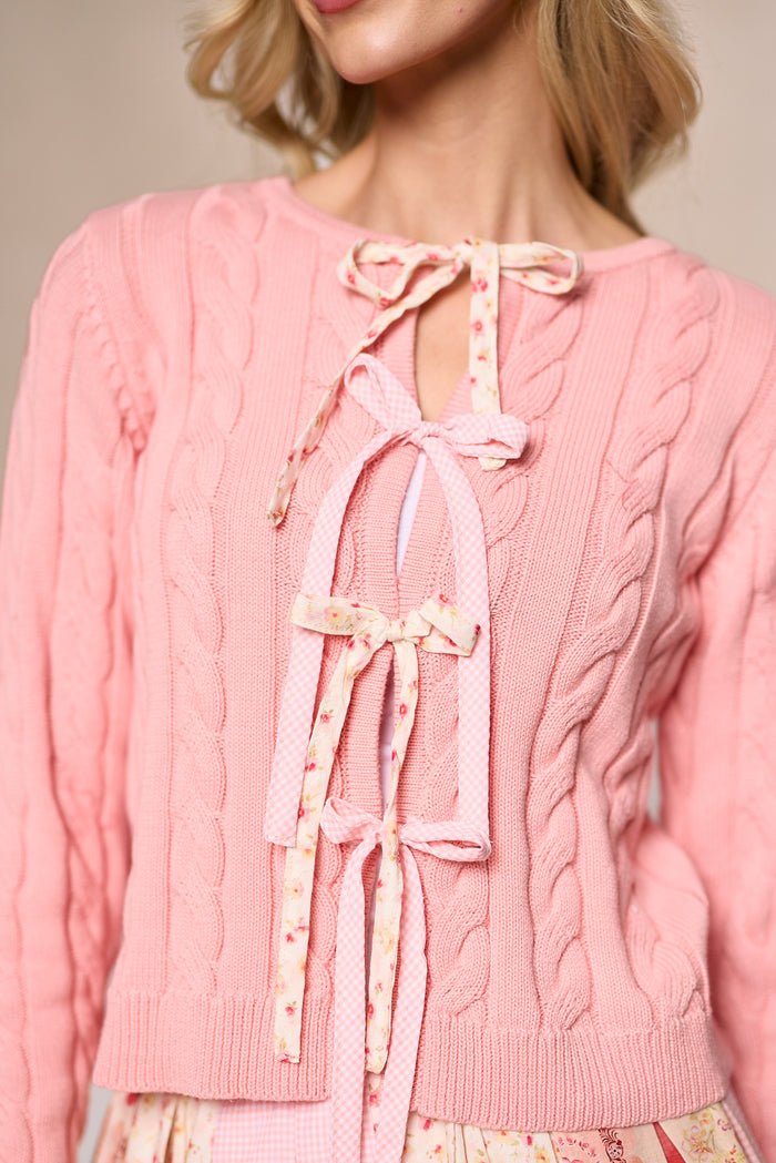 A woman wears the Imani Sweater in Blush by Ivy City Co, featuring three floral-patterned bows on the front, paired with a floral skirt. Only her neck and part of her face are visible.