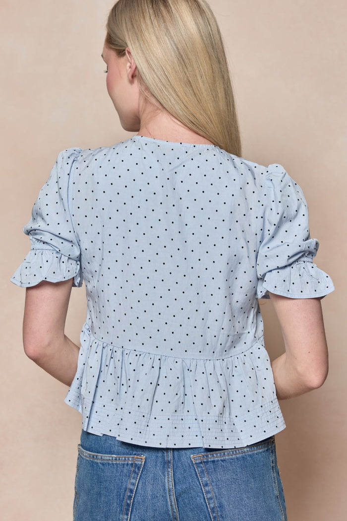 A woman with long blonde hair faces away, wearing the Ivy City Co Lavinia Top in Light Blue—a short-sleeved polka dot blouse featuring ruffled sleeves and a peplum hem—paired with blue jeans. Her hair is adorned with a delicate black lace bow, set against a neutral beige background.