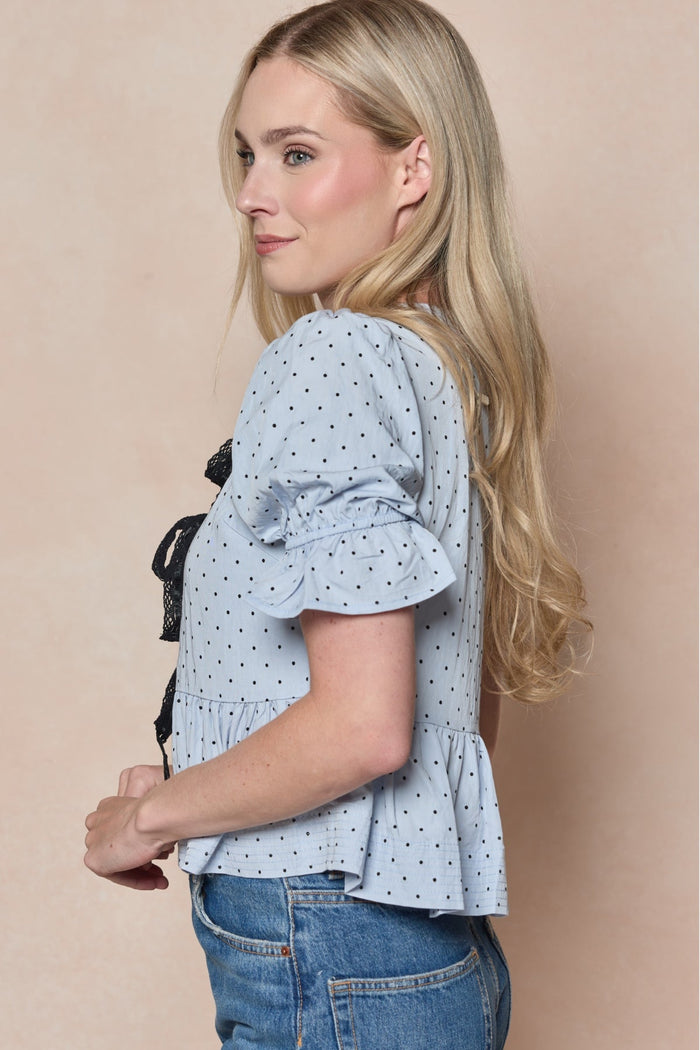 A woman with long blonde hair, styled with a delicate black lace bow, stands sideways against a beige background wearing Ivy City Co’s Lavinia Top in Light Blue—featuring polka dots and ruffled sleeves—paired with blue jeans.