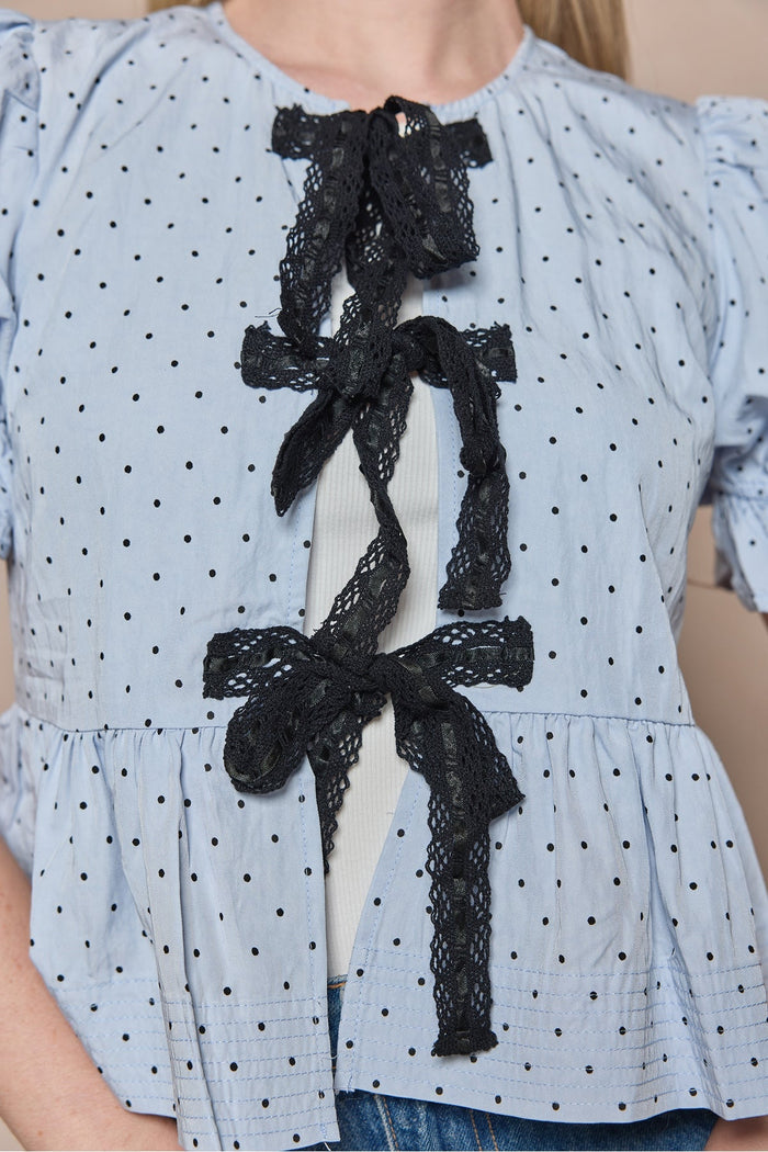 A person is shown from the torso up, wearing the Ivy City Co Lavinia Top in Light Blue—a short-sleeve blouse featuring playful polka dots and delicate black lace bow accents layered over a white top.