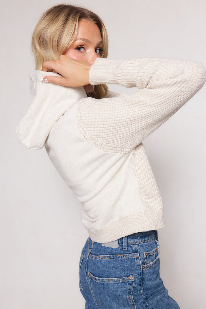 A woman with long blonde hair poses sideways against a plain background, wearing the Ivy City Co Lisbeth Hoodie in Oatmeal with textured sleeves and blue jeans. She lifts one arm to her face, highlighting the hoodie’s relaxed silhouette.