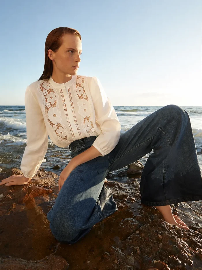Woman sitting on a rocky beach wearing a white blouse with lace details and blue jeans.