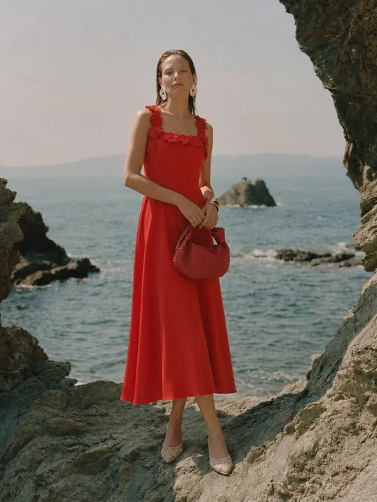A woman stands on rocky coastal ground wearing a bright red midi dress with ruffled shoulder straps, holding a matching red handbag, with the ocean and sea stacks in the background.
