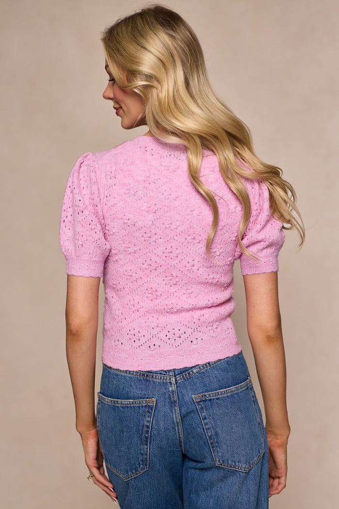 A woman with long blonde hair, seen from behind, wears the Ivy City Co Michaela Cardigan in Pink—a light pink cropped jacket with subtle eyelet details and a fitted button-front—paired with blue high-waisted jeans, against a neutral background.