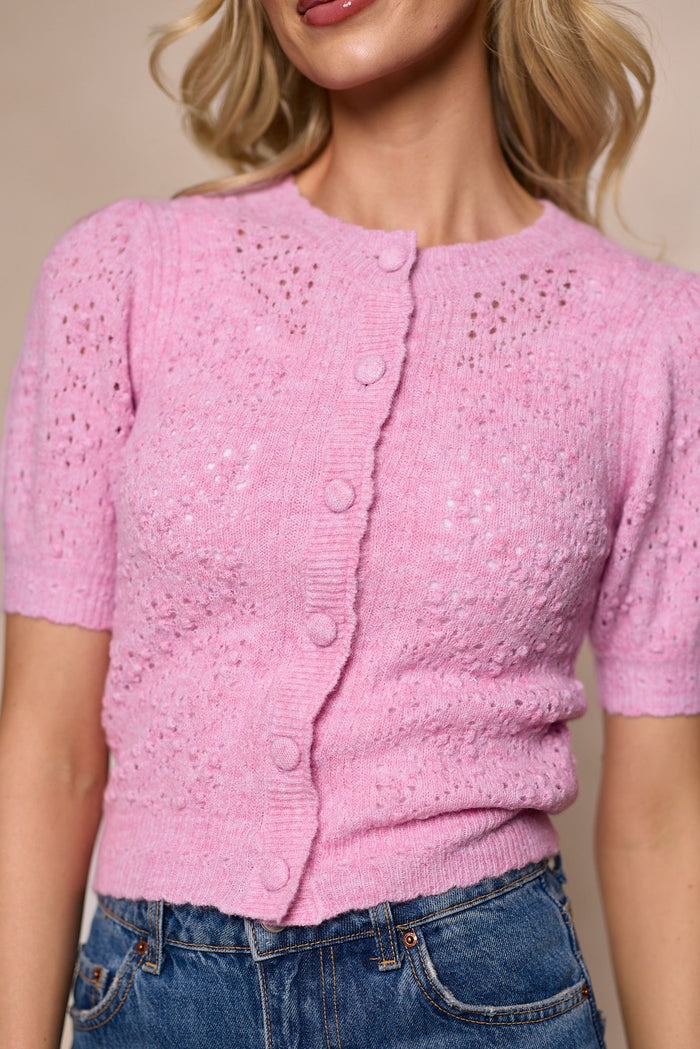 A woman wearing the Michaela Cardigan in Pink by Ivy City Co—a light pink, short-sleeve, fitted button-front knitted sweater with eyelet details—paired with high-waisted blue jeans.