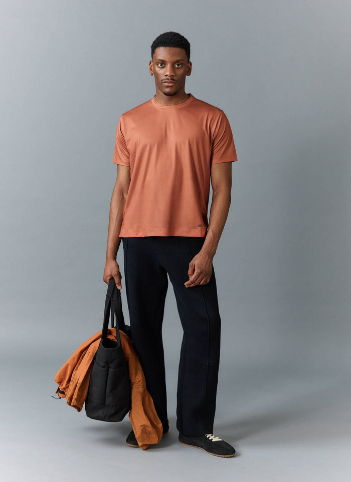 A model wears the Percival PCVL Performance Top in Rust, styled with black trousers and carrying a black bag and jacket.