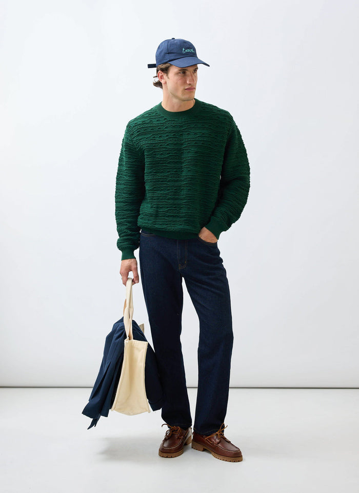 A model wears the Percival Tales Knitted Crew Neck Jumper in forest green styled with dark jeans, a blue cap, and a tote bag.